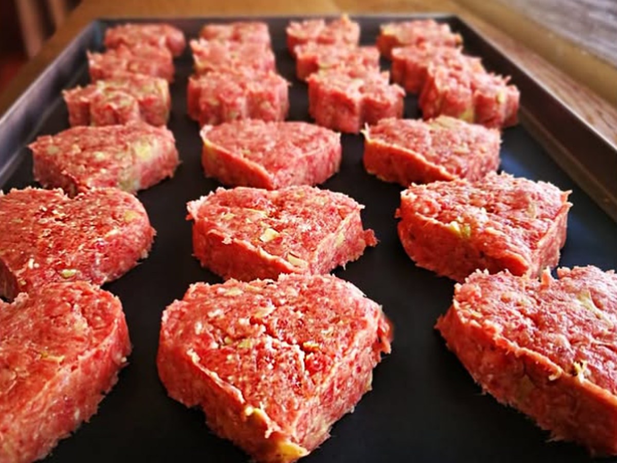 Medaglioni di carne macinata a forma di cuore con pezzetti chiari, crudi su una teglia scura appoggiata su legno